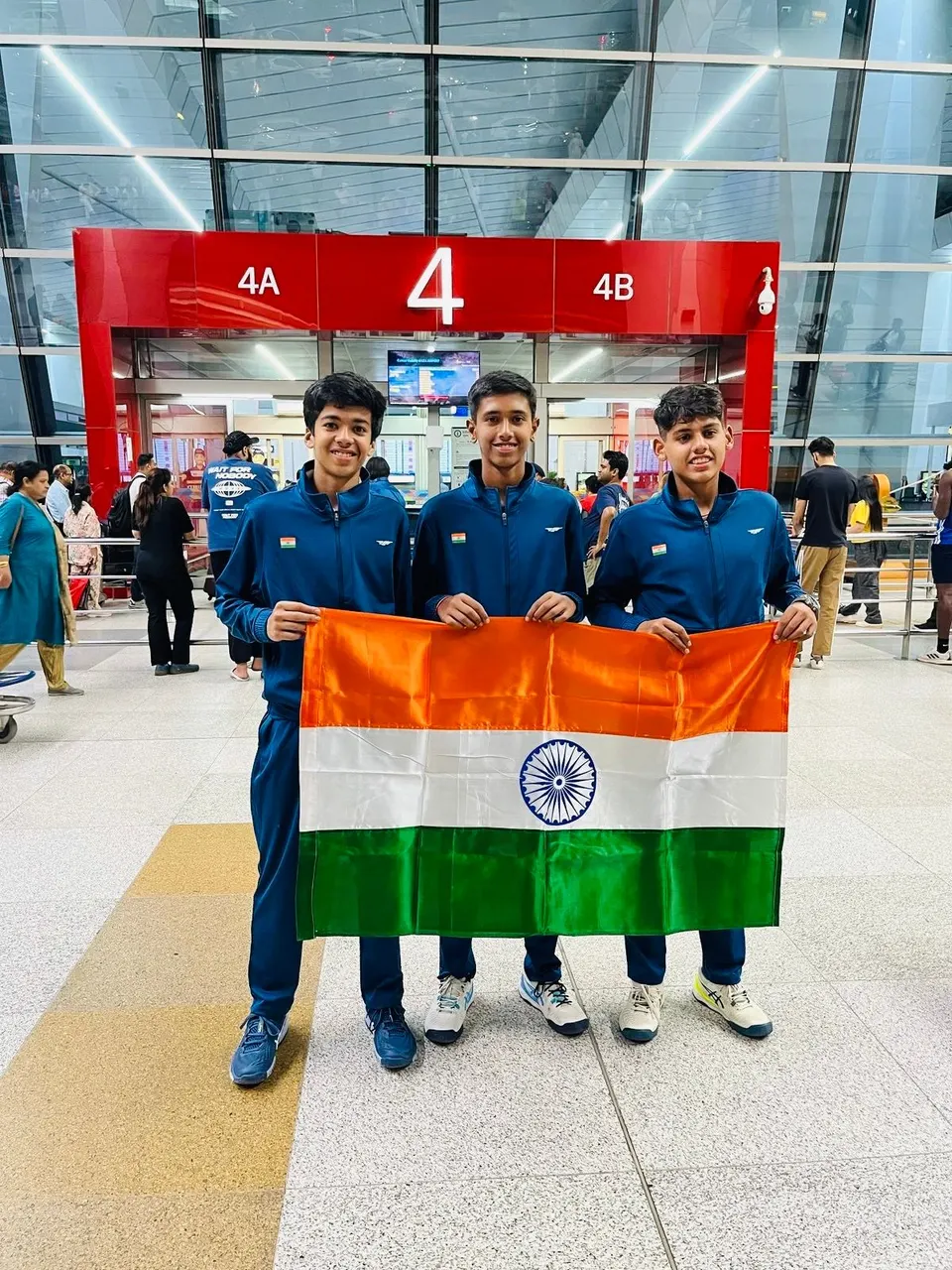 Three junior players in India team tracksuits holding the Indian flag at an airport